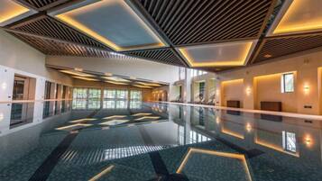 A spacious indoor swimming pool with a modern design, featuring geometric patterns on the floor and ceiling, and a long corridor with windows on one side.