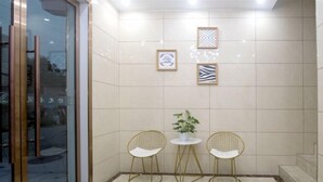 Un área de espera pequeña y moderna con dos sillas de alambre dorado, una mesita, una planta en maceta y cuadros en la pared.