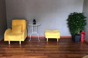 Une pièce avec un fauteuil jaune, une petite table ronde, un pouf assorti, une plante en pot et un objet rouge posé sur le sol.