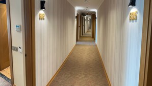 Un corridoio d'albergo con pavimento rivestito di moquette, porte in legno e lampade a parete.