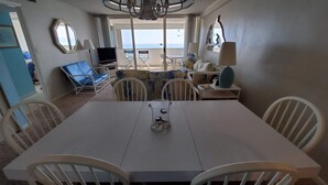 Un comedor con una mesa y sillas blancas, un televisor y vista al mar.