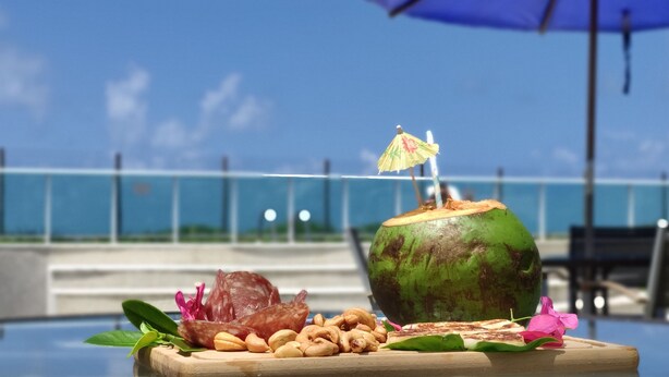 A wooden table with a coconut, sliced meat, nuts, and a small umbrella on a sunny day by a pool with a clear view of the ocean. - Pé na Areia Perfeito para 7 Pessoas + Garagem (Barra de São Miguel)