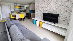 A modern living room with a grey sofa, a flat-screen TV on a white stand, and a kitchen in the background. - Pé na Areia Perfeito para 7 Pessoas + Garagem (Barra de São Miguel)