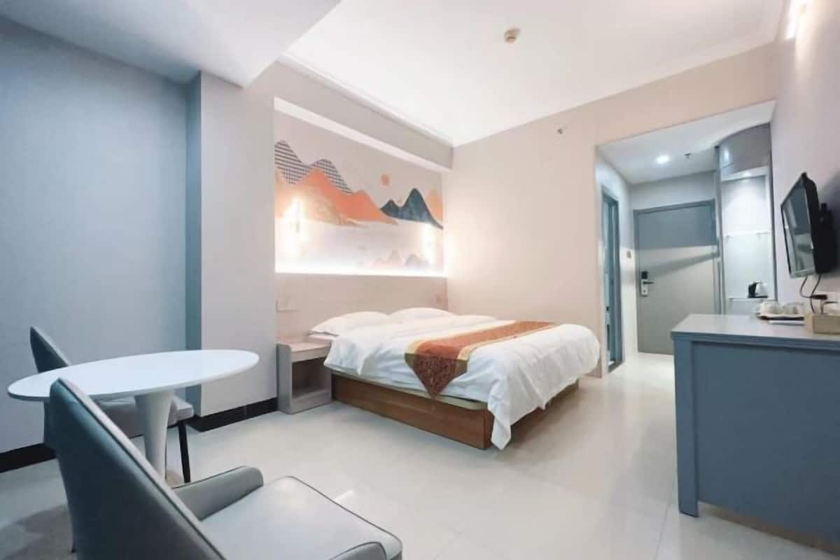 A modern hotel room with a bed, a desk, a chair, a small table, and a wall-mounted TV.