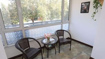 Una habitació amb un finestral gran, dues cadires de vimet, una taula petita rodona amb un ram de flors i un quadre amb marc a la paret.