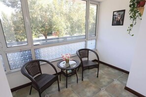 Una habitación con un ventanal grande, dos sillas de mimbre, una mesita redonda con un jarrón de flores y un cuadro enmarcado en la pared.