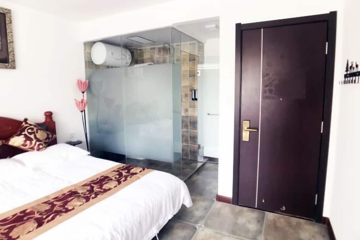 A hotel room with a bed, a dark wooden door, and a shower area with glass doors.