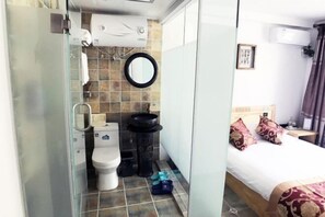 A bathroom with a toilet, bidet, and a mirror. There is a bed with pillows and a bedside table with a lamp. - Fushun ala warm hotel (Fushun)