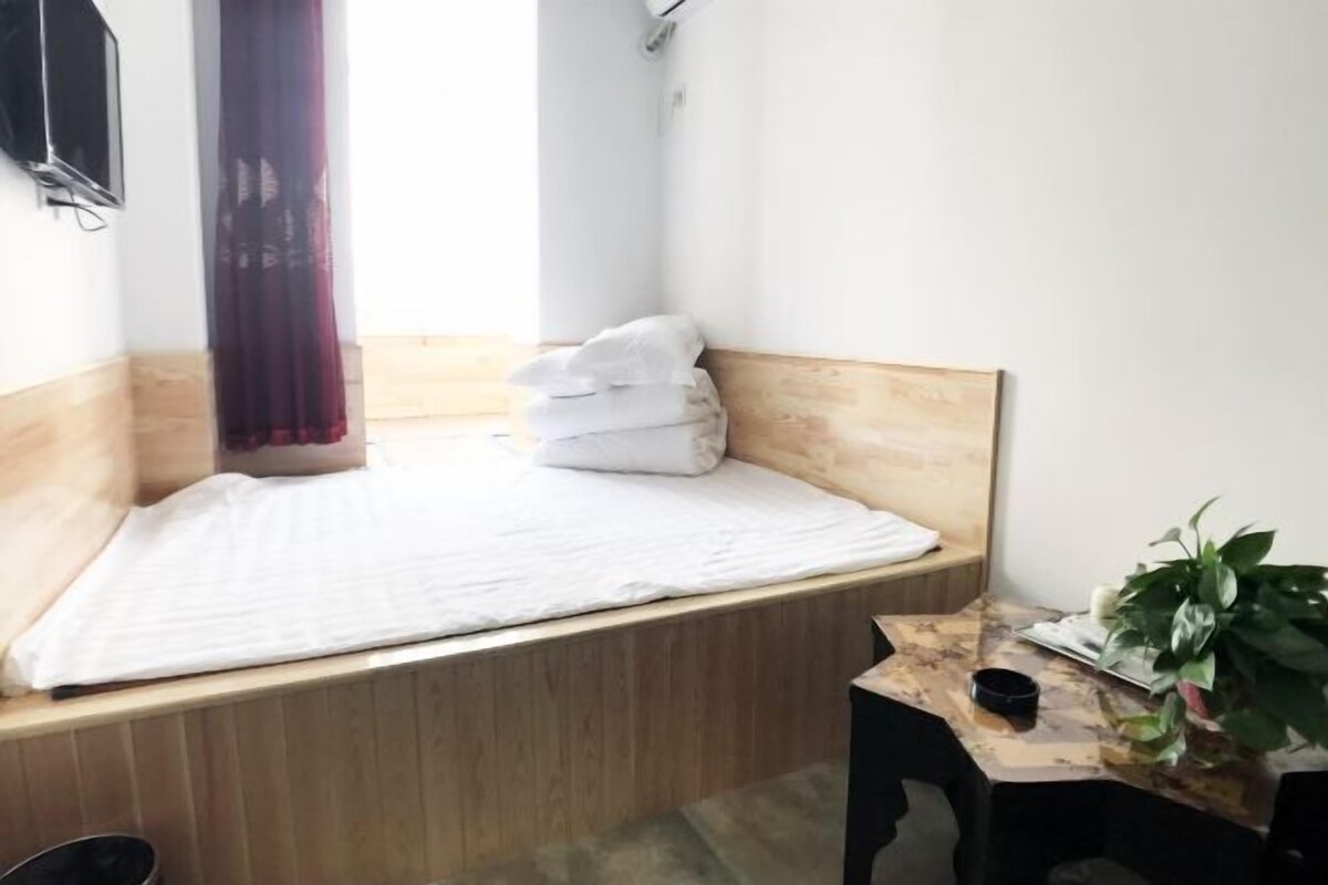 A small, neatly arranged room with a bed, a small table with a plant, and a window with curtains.
