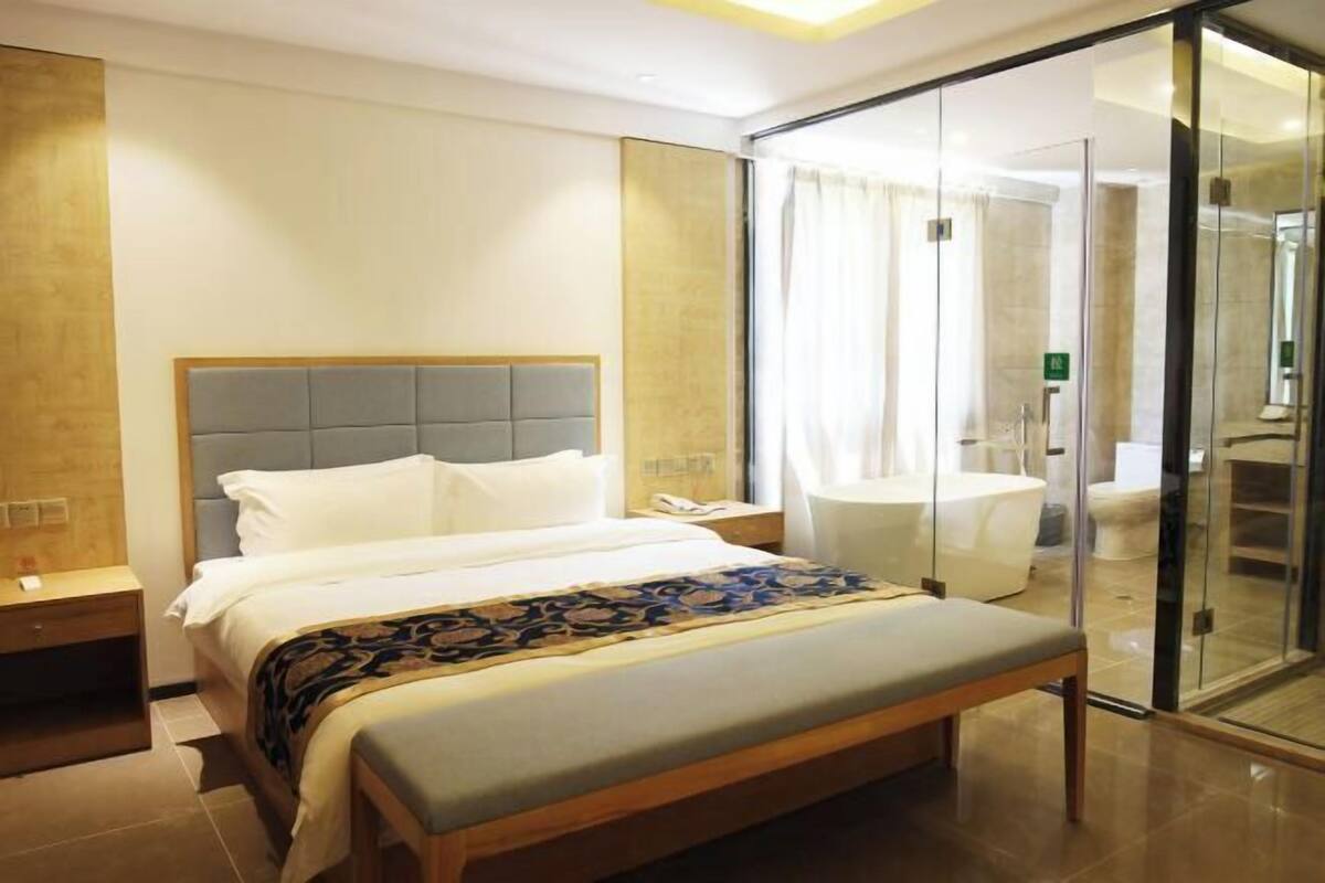 Una habitación de hotel moderna con una cama grande, una mesita de noche, una zona de ducha con mampara de vidrio y un pequeño rincón de descanso.