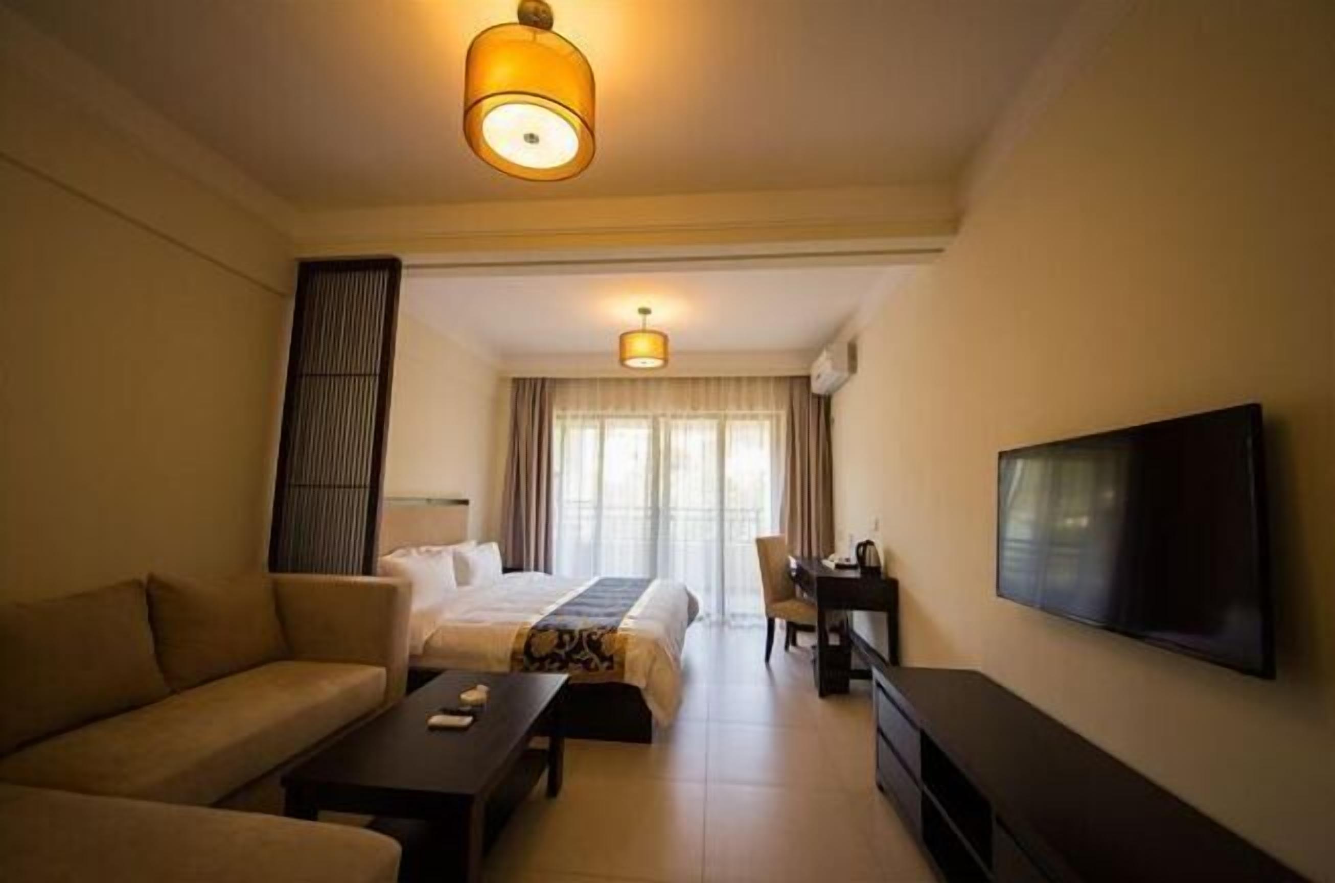 Habitación de hotel con cama, sofá, televisor, escritorio y un ventanal con cortinas.