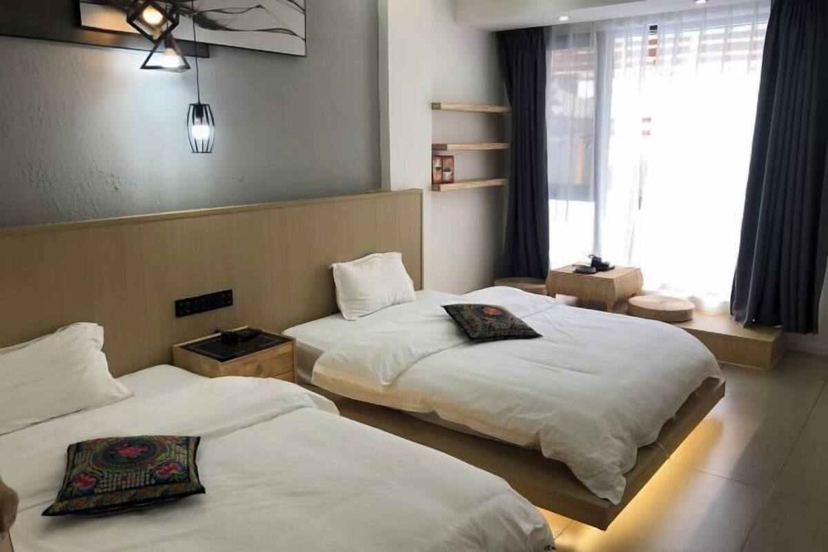 A hotel room with two beds, a bedside table, a lamp, a window with curtains, and a shelf with a picture frame.