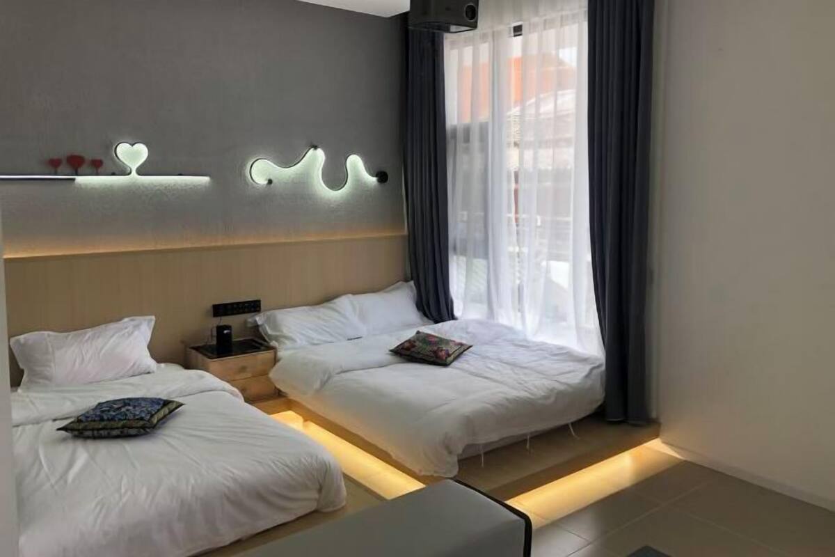 A hotel room with two beds, a bedside table with a lamp, a wall-mounted shelf with decorative items, and a window with curtains.