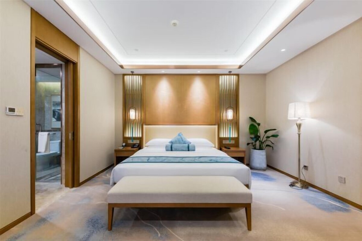 A modern hotel room with a large bed, bedside tables, a bench, a potted plant, and a floor lamp.