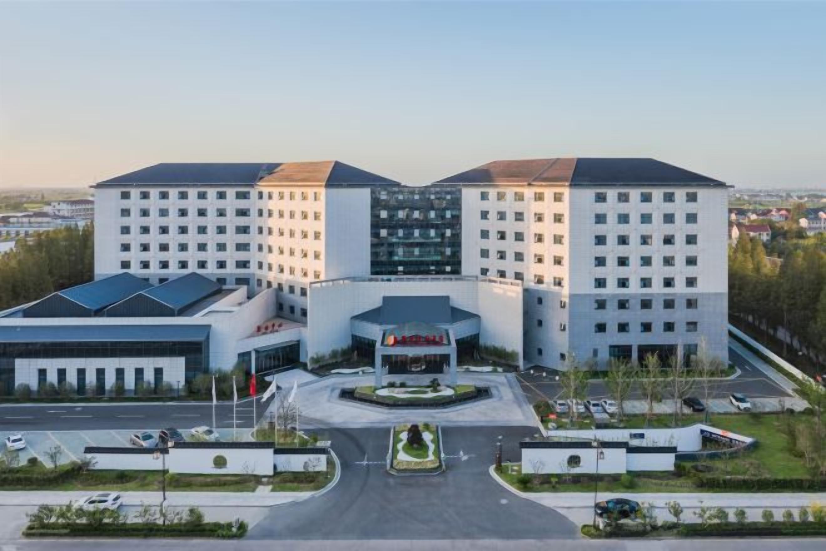 Un hotel de varios pisos con entrada central, zona de estacionamiento y jardín ajardinado.