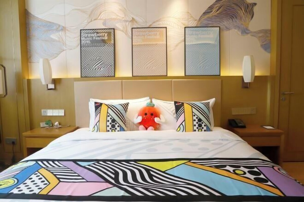 A hotel room with a bed, pillows, a stuffed toy, a wall with framed pictures, and a nightstand.