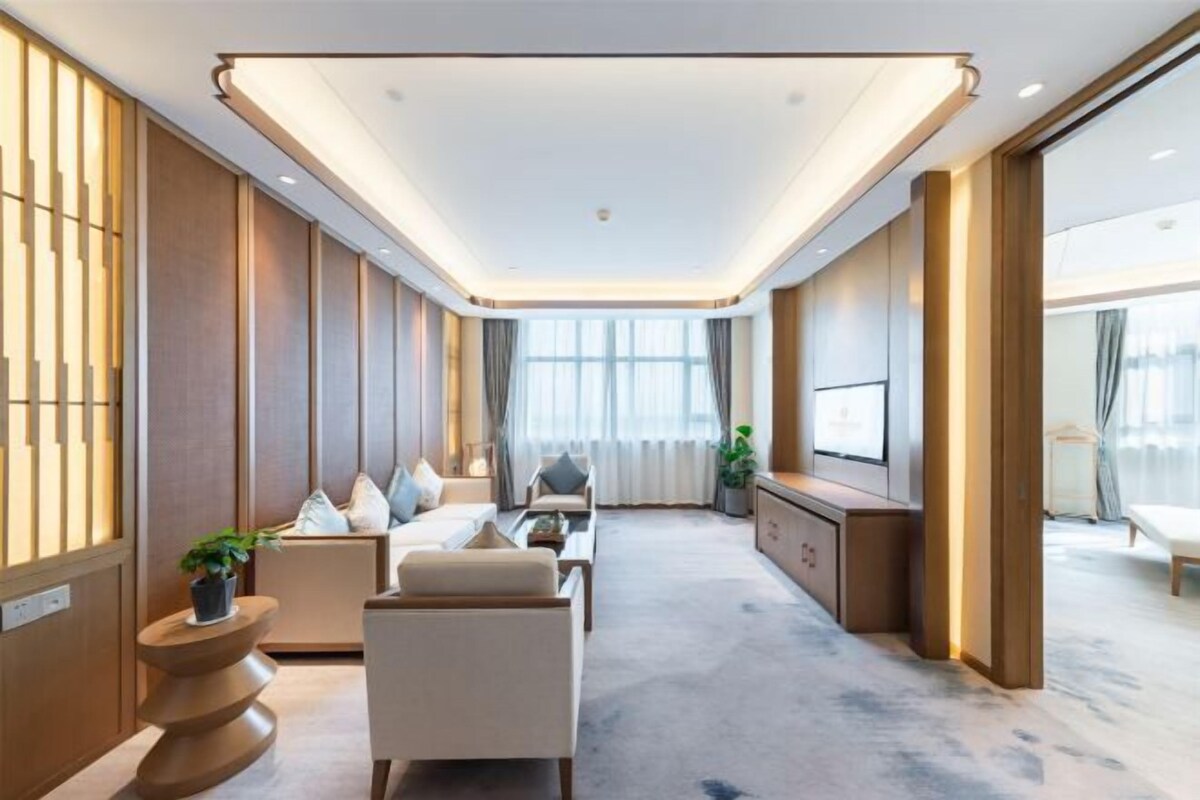 Una habitación de hotel moderna con un sofá, sillones, un televisor y un ventanal grande.