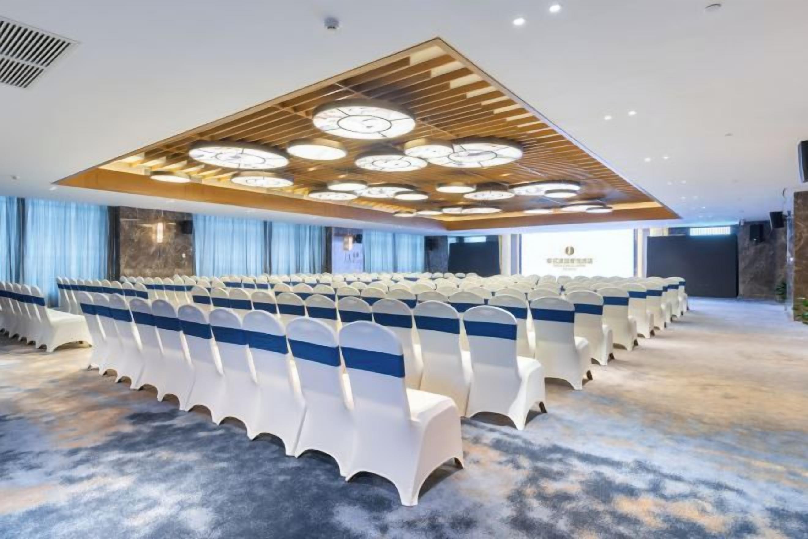 Uma sala de conferências com fileiras de cadeiras, detalhe no teto de madeira e piso coberto de carpete.