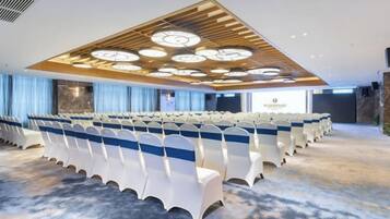Uma sala de conferências com fileiras de cadeiras, detalhe no teto de madeira e piso coberto de carpete.
