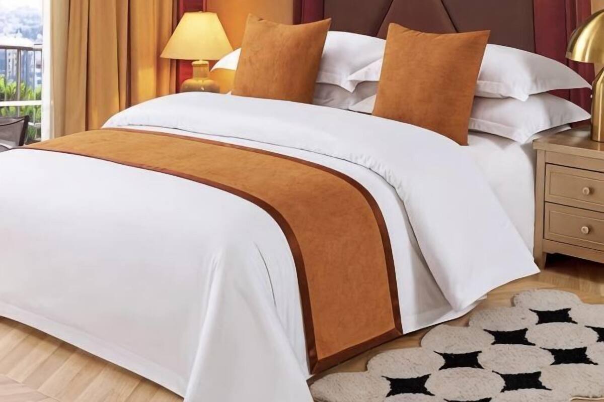 Una cama bien hecha con cubrecama blanco y marrón, una mesita de noche de madera con una lámpara, y una alfombra estampada en el suelo.