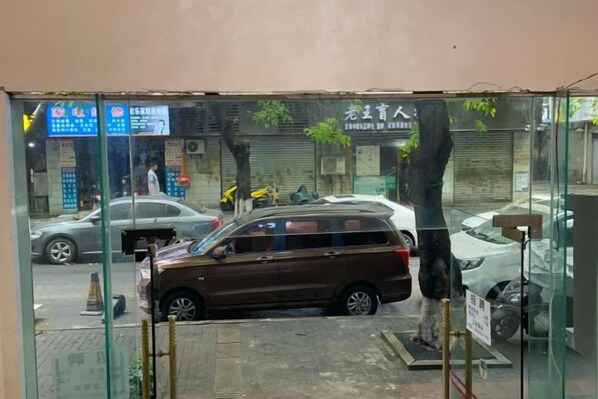 A view through a glass door of a street with parked cars, a signboard, and a building with a closed shutter.