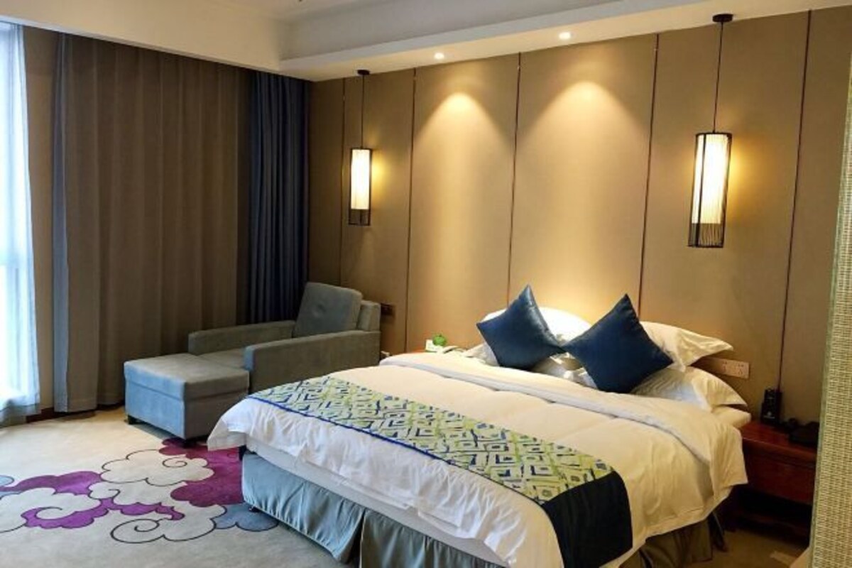 A hotel room with a bed, a chair, a nightstand, and two wall-mounted lamps.