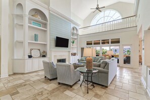 A spacious living area with a high ceiling, a fireplace, a television, and a balcony with a view.