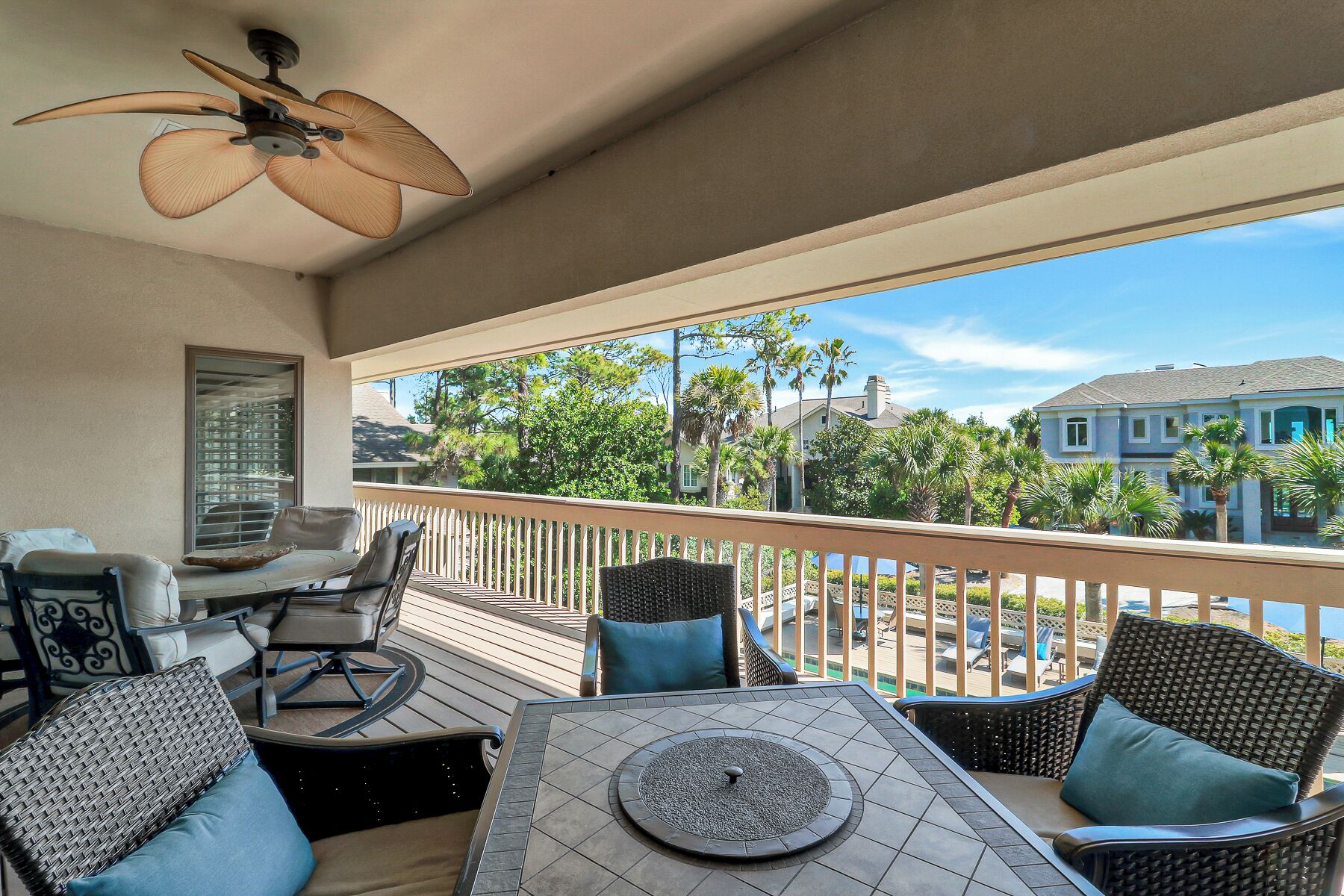A covered outdoor area with a table and chairs, a ceiling fan, and a view of a residential area with palm trees and houses.