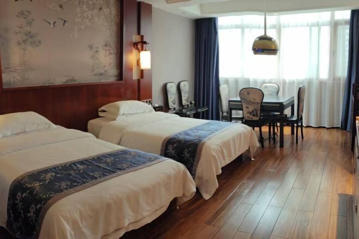 A hotel room with two beds, a desk with a chair, a large window with curtains, and a wall with a mural.