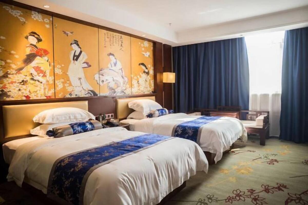 A hotel room with two beds, a traditional painting on the wall, a window with curtains, and a floral carpet.