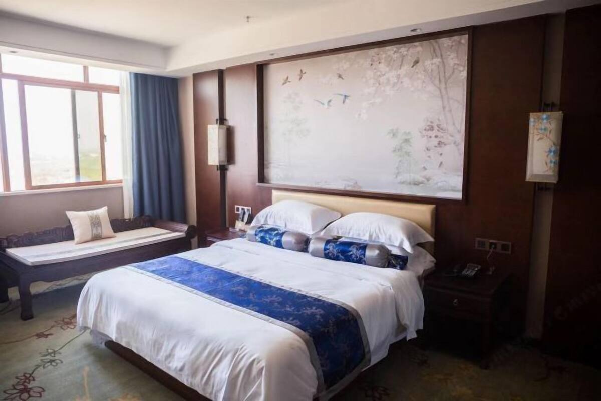 A hotel room with a large bed, a bedside table, a nightstand, a window with curtains, and a wall-mounted artwork.