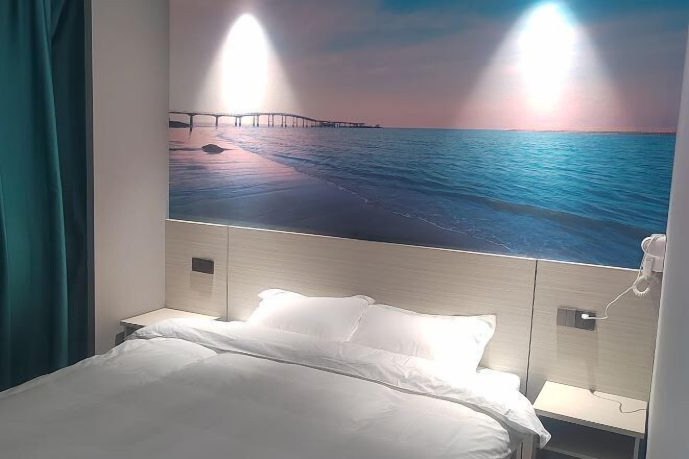 Ein Hotelzimmer mit einem Bett, einem Nachttisch, einem an der Wand montierten Telefon und einem Gemälde eines Piers über dem Wasser.