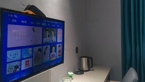 A room with a flat-screen TV mounted on the wall displaying various content, a kettle, a small table, and a chair. - JYhotel (Yuyao)