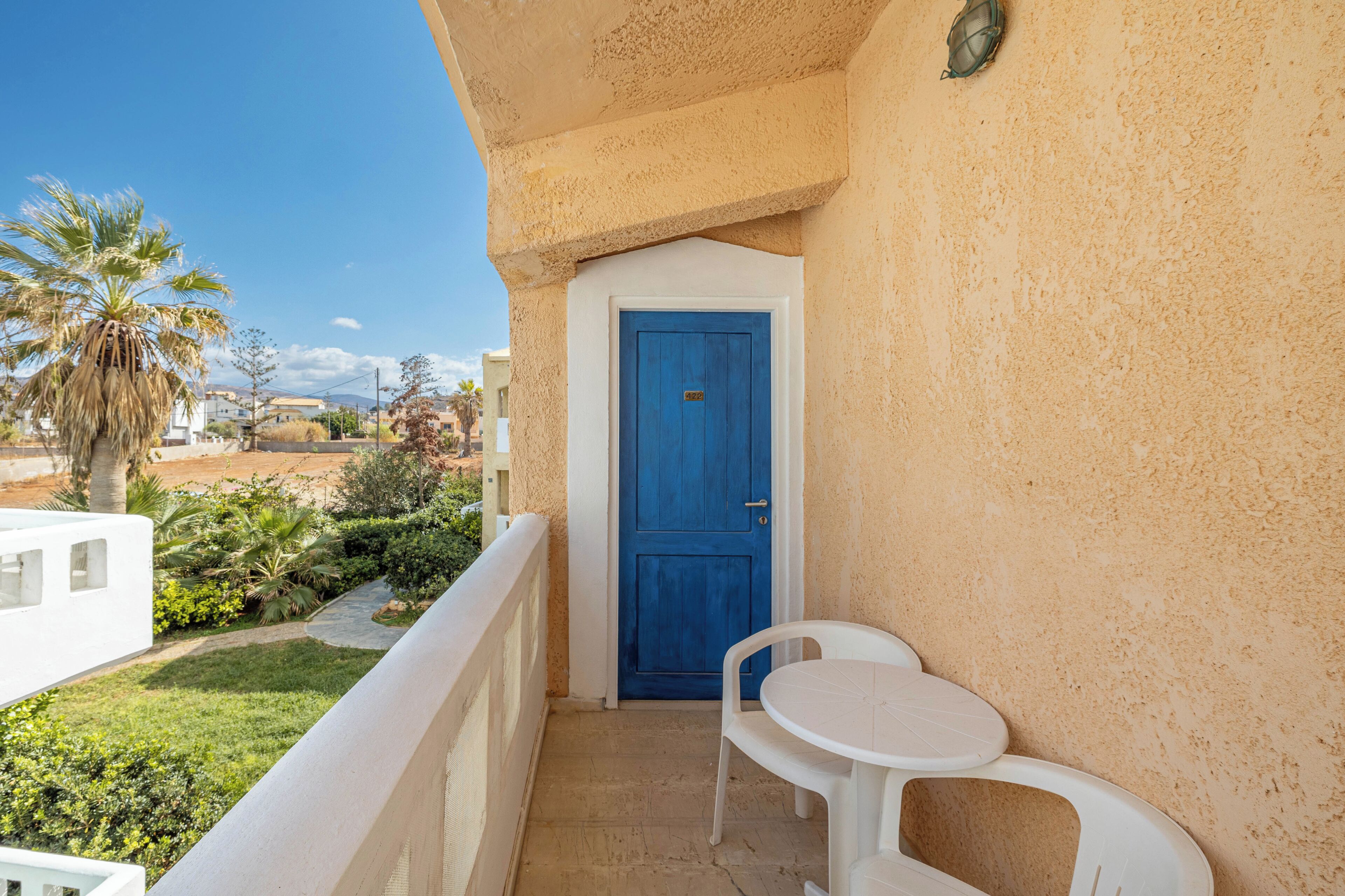 Un balcón con una puerta azul, sillas blancas y vistas a zonas verdes y edificios.
