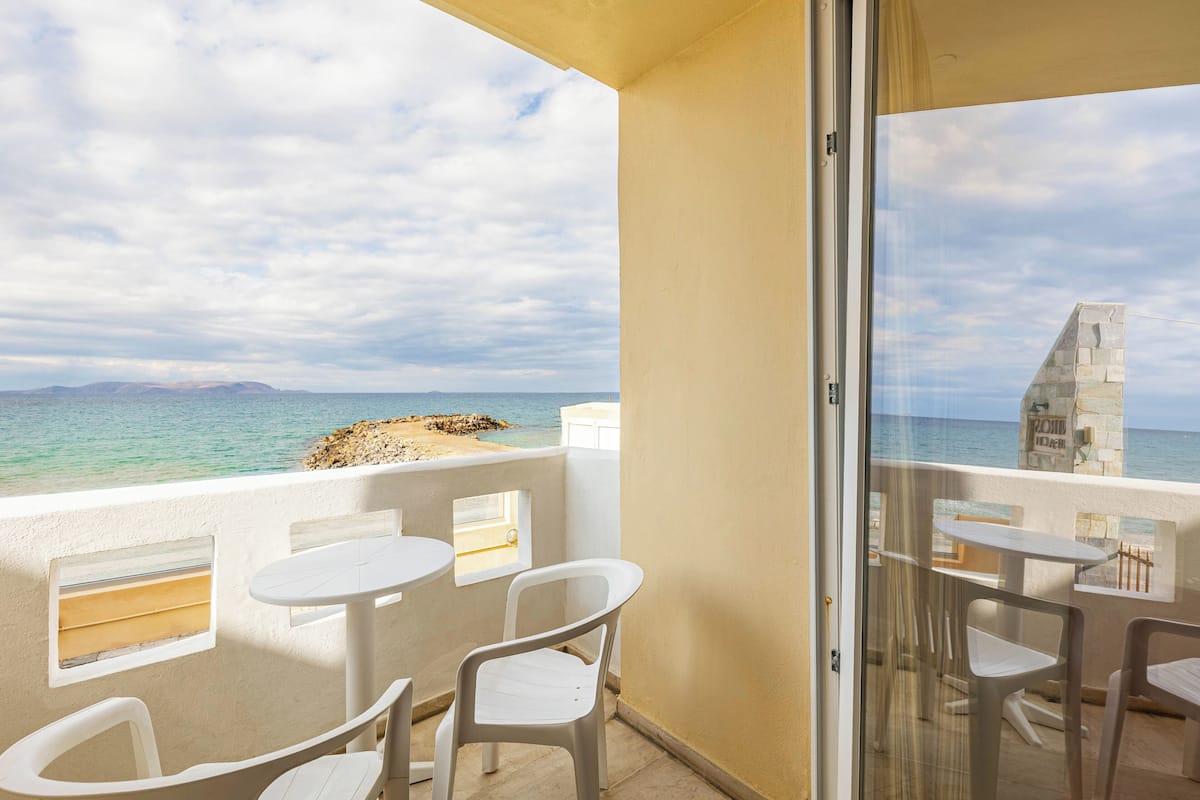 A balcony with white plastic chairs and tables, overlooking a beach and the sea.