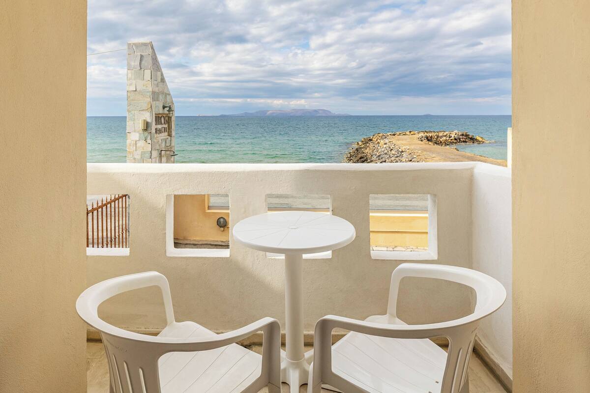A balcony with a white table and two chairs, overlooking a coastal view with a stone structure and a rocky shoreline.