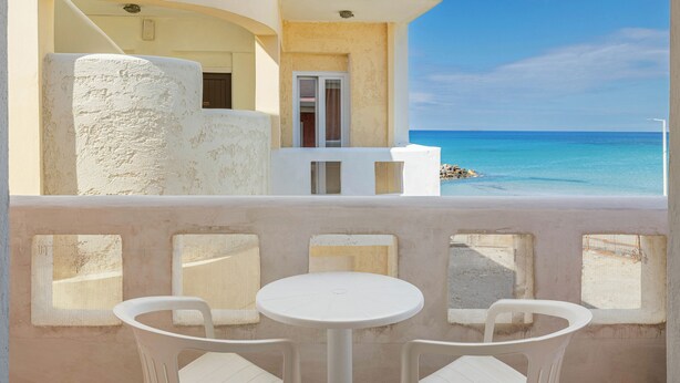 A balcony with white chairs and a table, overlooking a clear blue sea and a white building with a door.