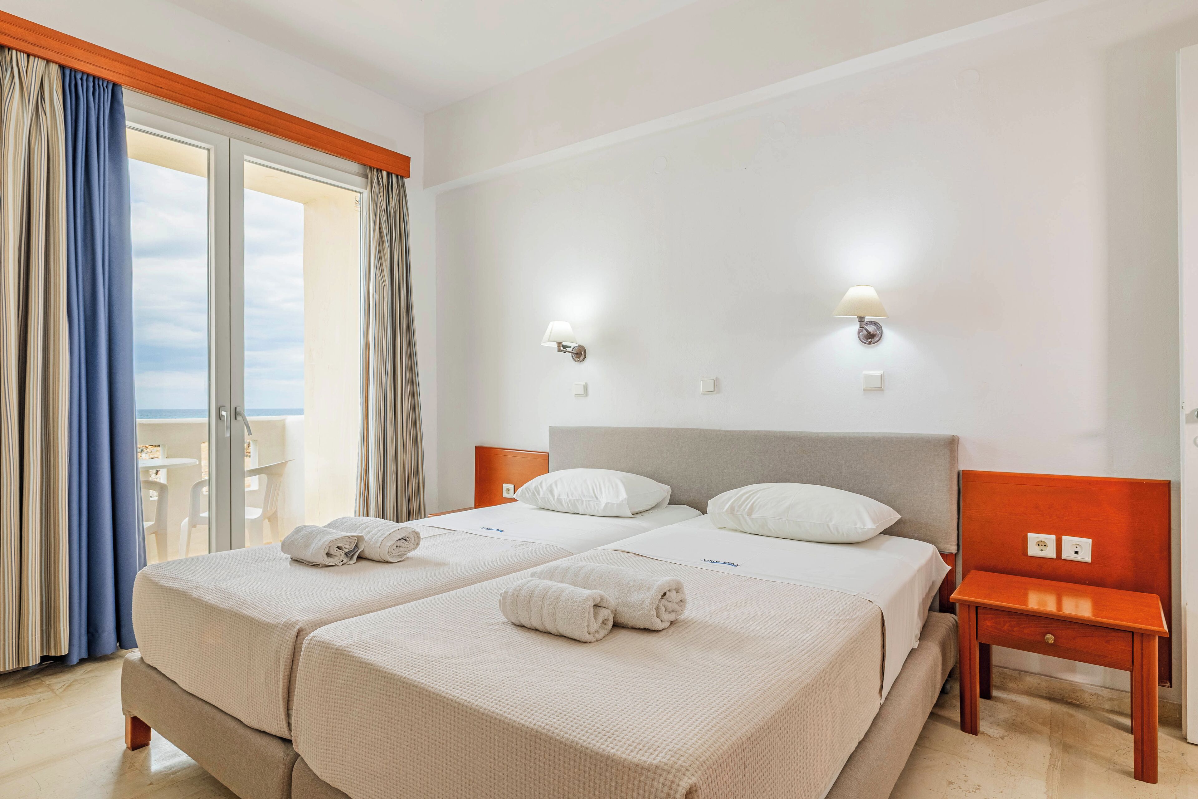 A hotel room with two beds, a wooden nightstand, and a view of the sea through the window.