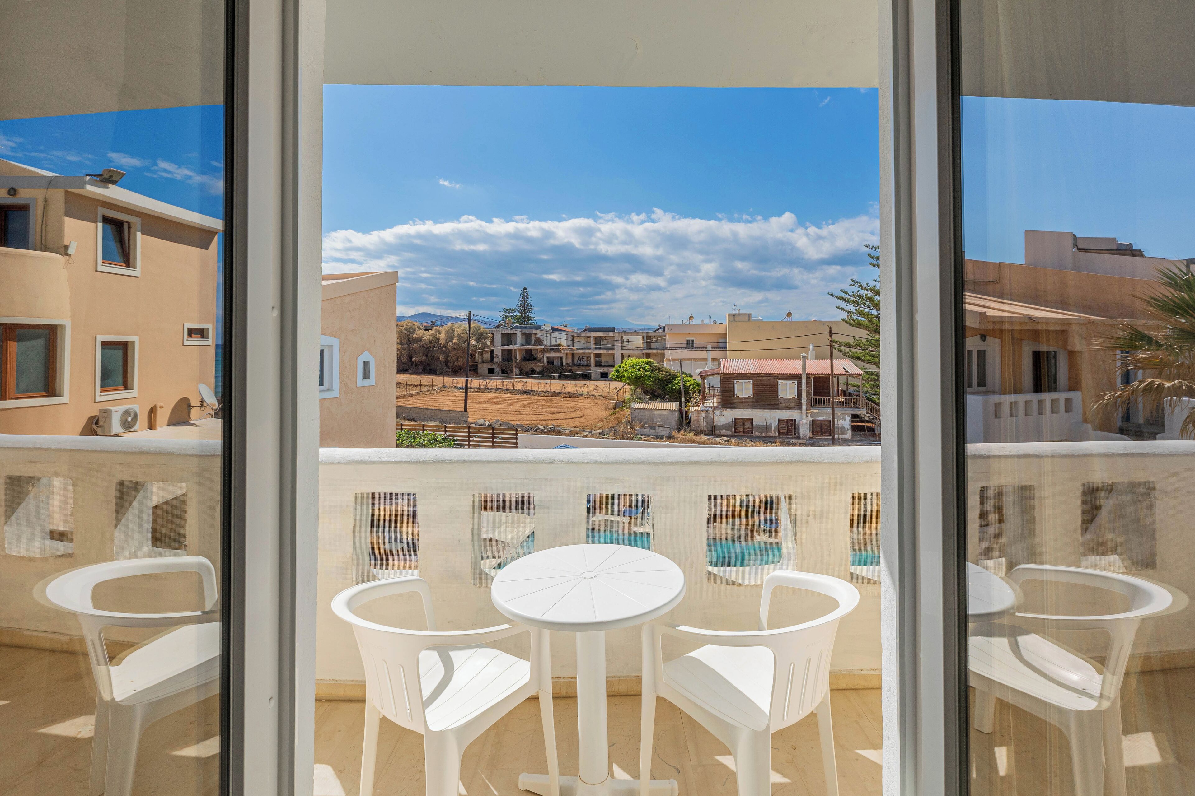 A balcony with white plastic chairs and a small round table, overlooking a residential area with houses and a clear sky.