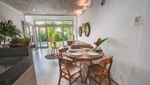 Un coin repas avec une table ronde dressée pour quatre personnes, entourée de chaises en bois. Une grande fenêtre offre une vue sur un jardin tropical.