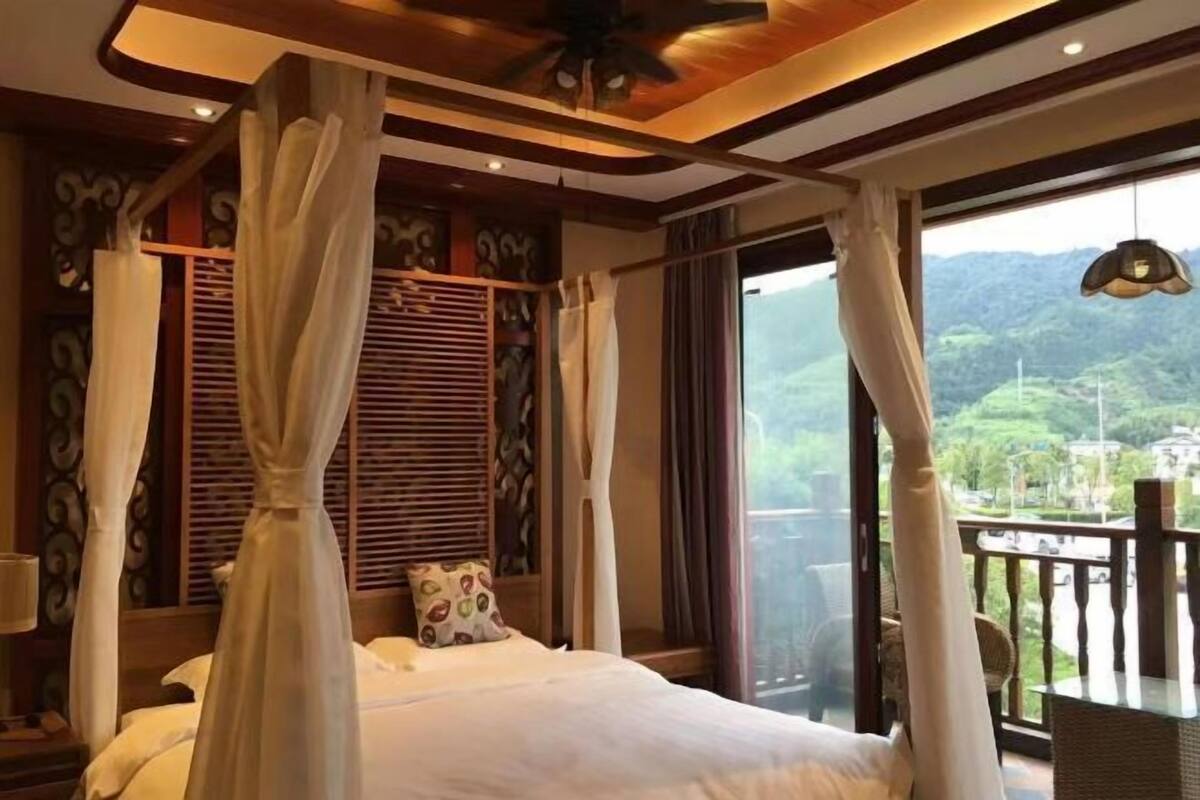 A bedroom with a bed, curtains, a ceiling fan, and a view of greenery outside.