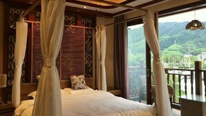 A bedroom with a four-poster bed, a balcony with a view of greenery, and a ceiling fan.