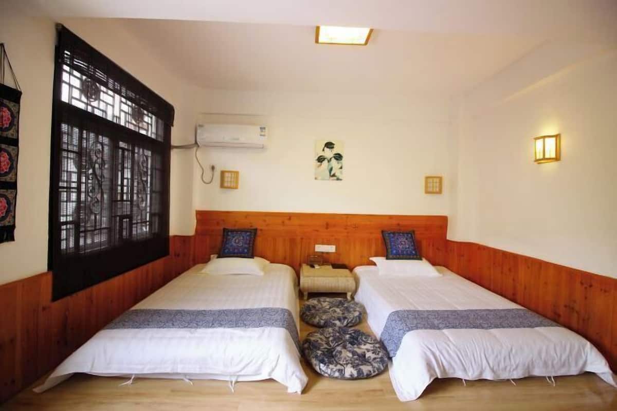 A room with two beds, a wooden headboard, a window with a decorative screen, an air conditioning unit, and a wall-mounted light fixture.