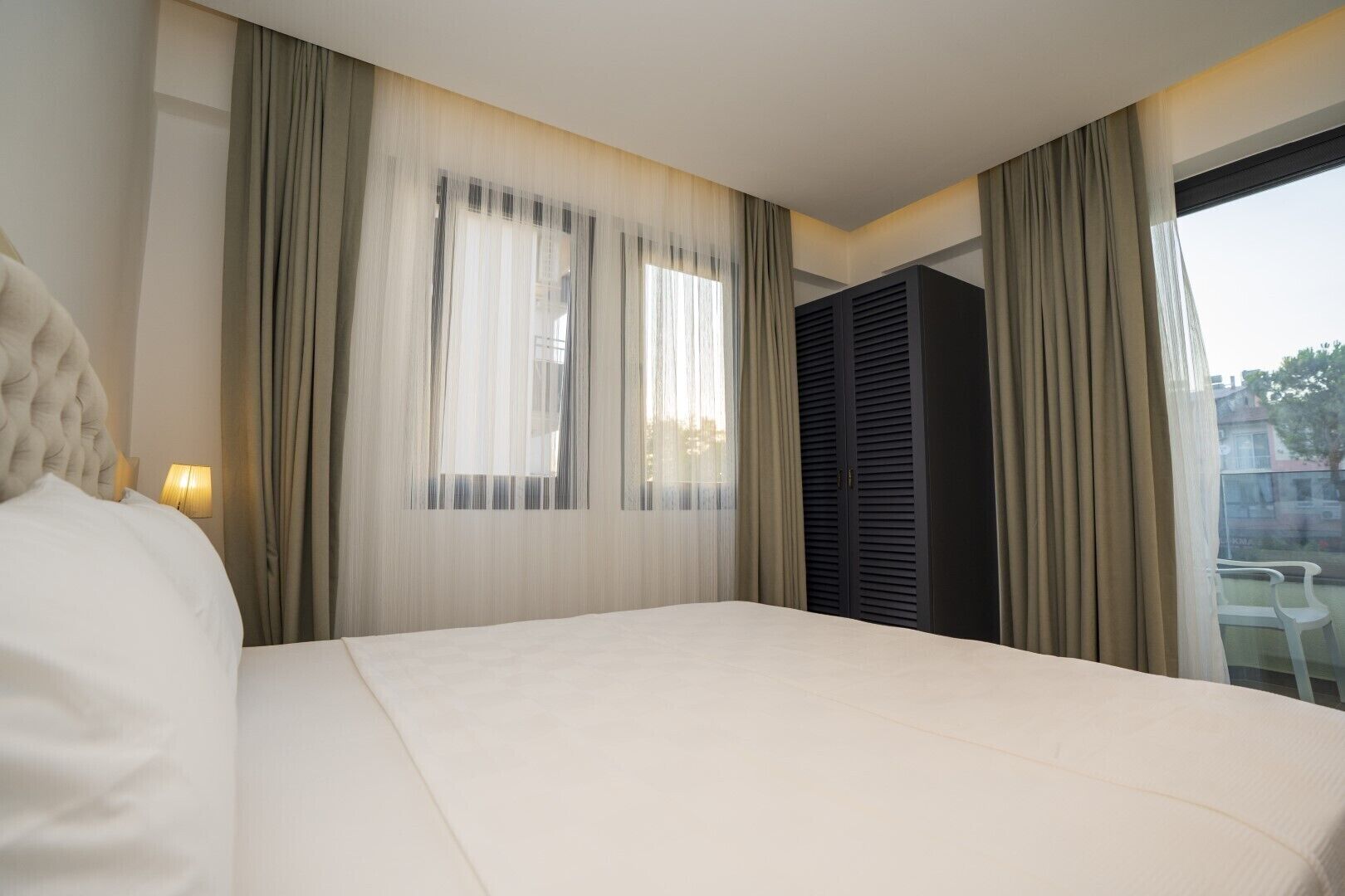 Quarto de hotel com uma cama grande, janela com cortinas leves, vista da cidade e varanda com mesa e cadeiras.