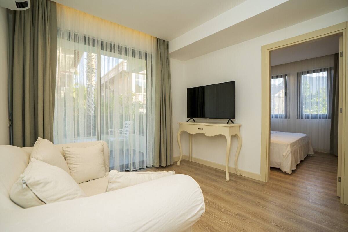 A hotel room with a large bed, a TV on a small table, and a balcony with sheer curtains.
