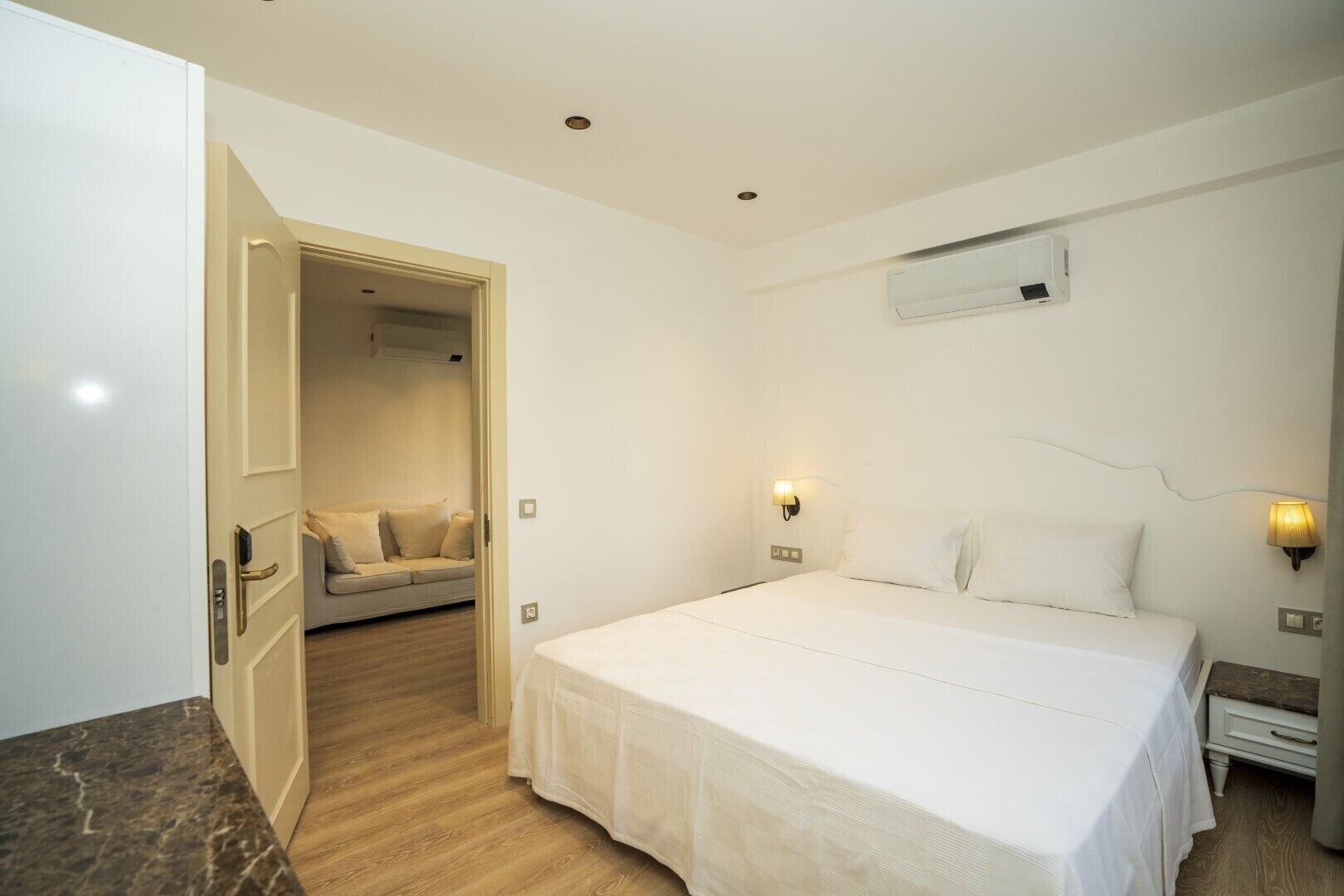 A hotel room with a large bed, a sofa in the adjacent room, and a wall-mounted air conditioner.