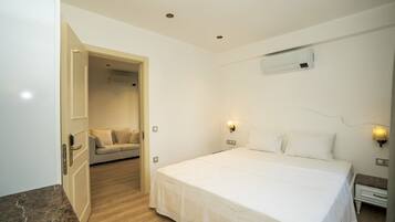 A hotel room with a large bed, a sofa in the adjacent room, and a wall-mounted air conditioner.