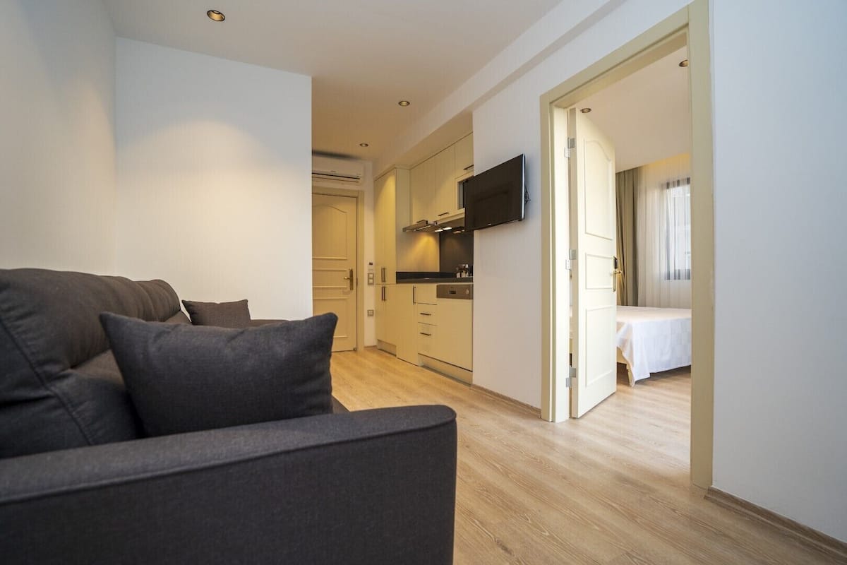 A compact living space with a sofa, a TV mounted on the wall, a kitchenette, and a bed in the adjacent room.