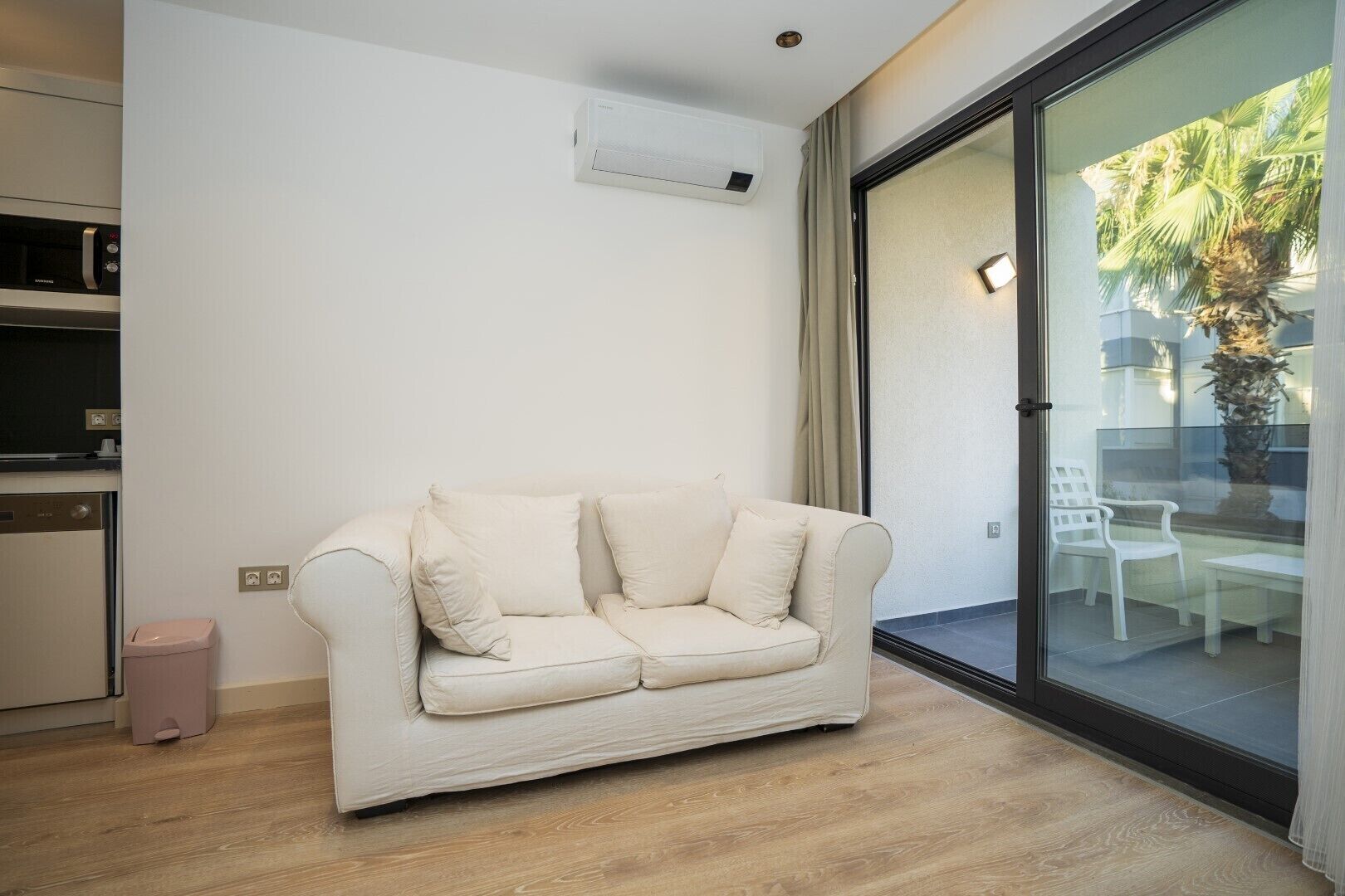 A modern living room with a beige sofa, a glass sliding door leading to an outdoor area with chairs and a palm tree, and a built-in air conditioning unit on the wall.