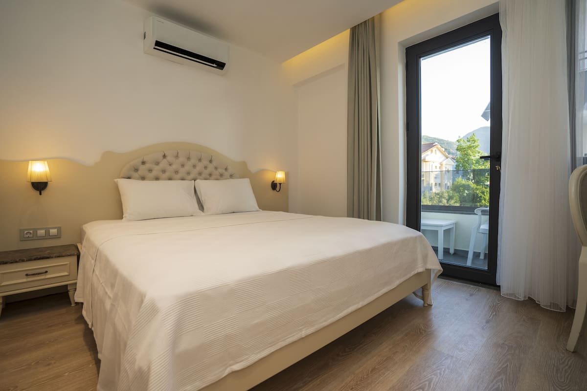 A neatly made bed with a headboard, two bedside lamps, a small bedside table, and a view of a balcony with a table and chairs.